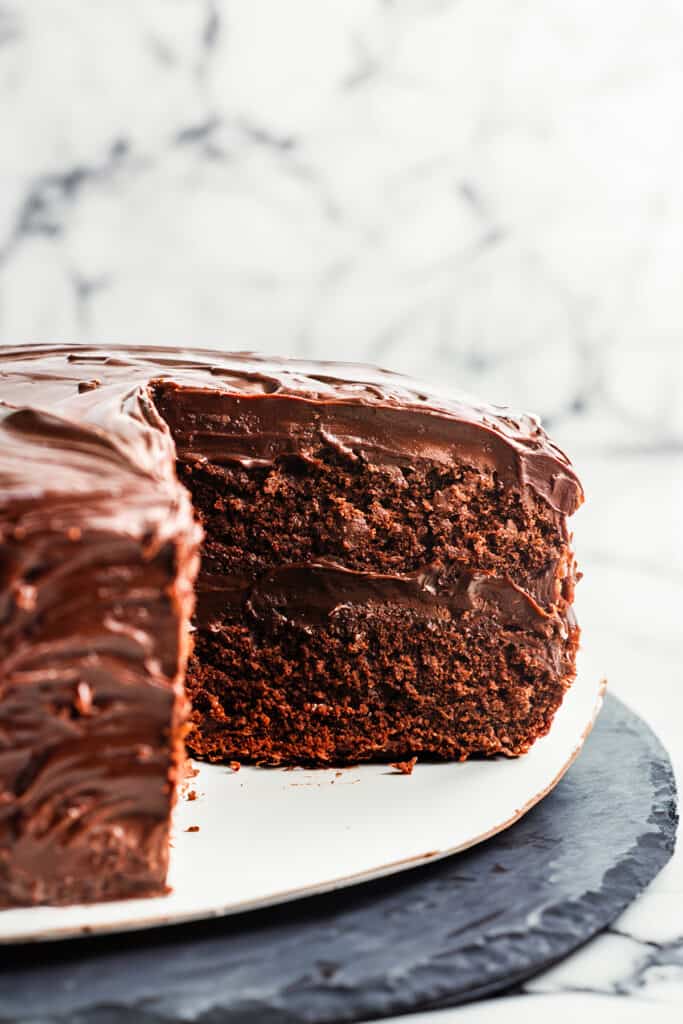 A devil's food cake with a slice removed, displaying the rich layers of cake and smooth chocolate frosting, on a white plate atop a dark slate board, against a marble background.