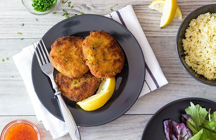 the crab cakes on a plate with lemon wedges and a dish of rice and a salad bowl in the background 