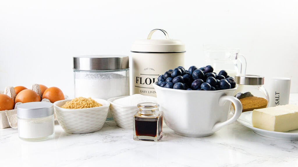 A variety of baking ingredients are arranged on a white countertop, ready to create Blueberry Crumb Cake Muffins. Eggs, flour, sugar, butter, blueberries, vanilla extract, salt, and milk sit neatly in containers and bowls.