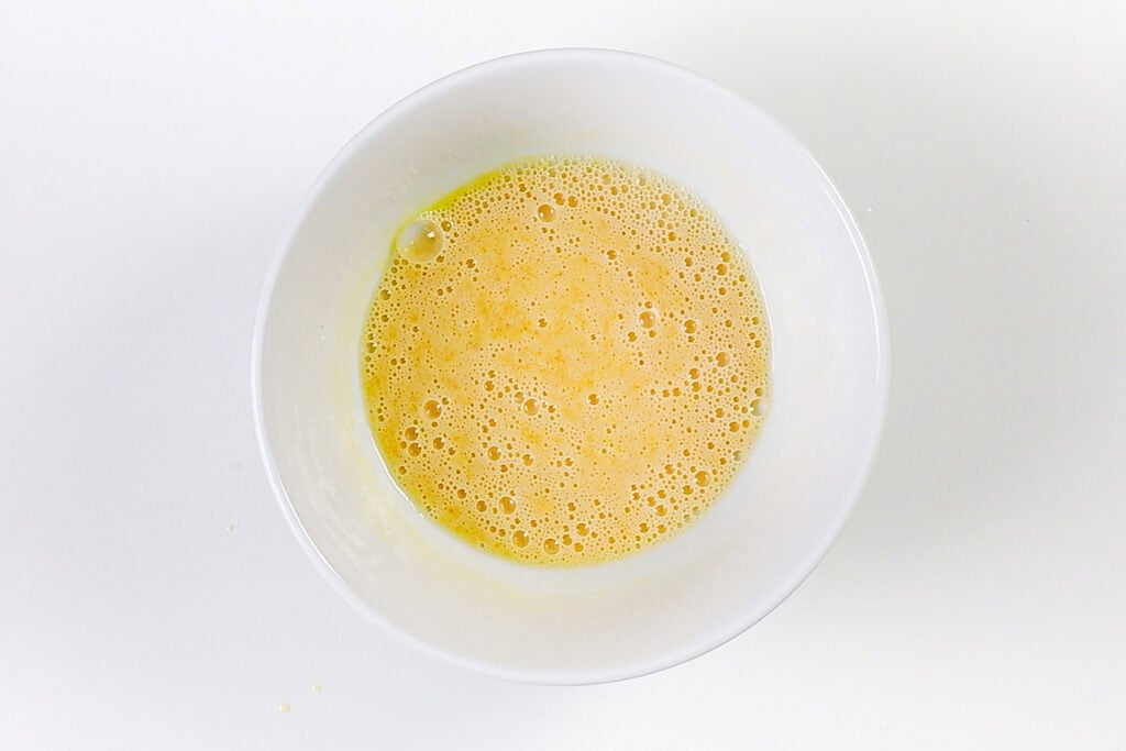 A white bowl filled with beaten eggs, creating a yellow, frothy mixture, sits on a white surface—perfect for preparing Blueberry Crumb Cake Muffins.