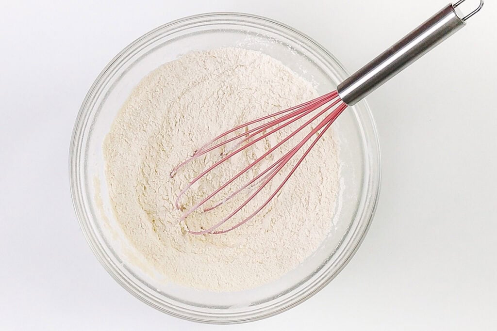 A glass bowl filled with flour and a pink-handled metal whisk resting inside, viewed from above on a white background—perfect for whipping up Blueberry Crumb Cake Muffins.