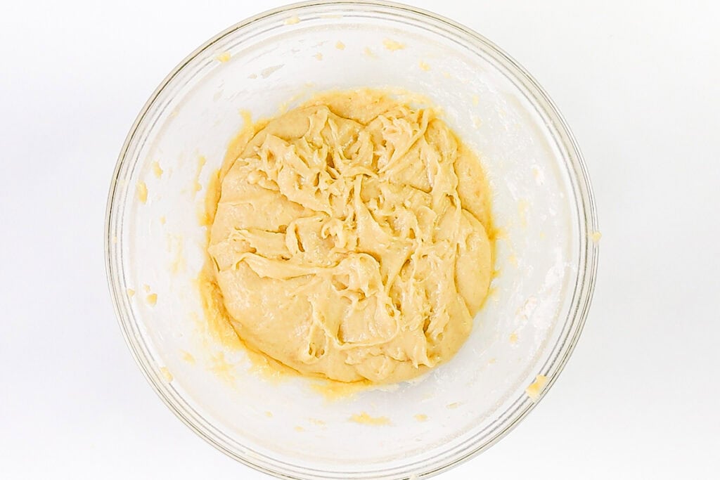 A glass bowl filled with smooth, pale yellow bread dough sits on a white surface, ready to rise or be shaped for baking delicious treats like Blueberry Crumb Cake Muffins.