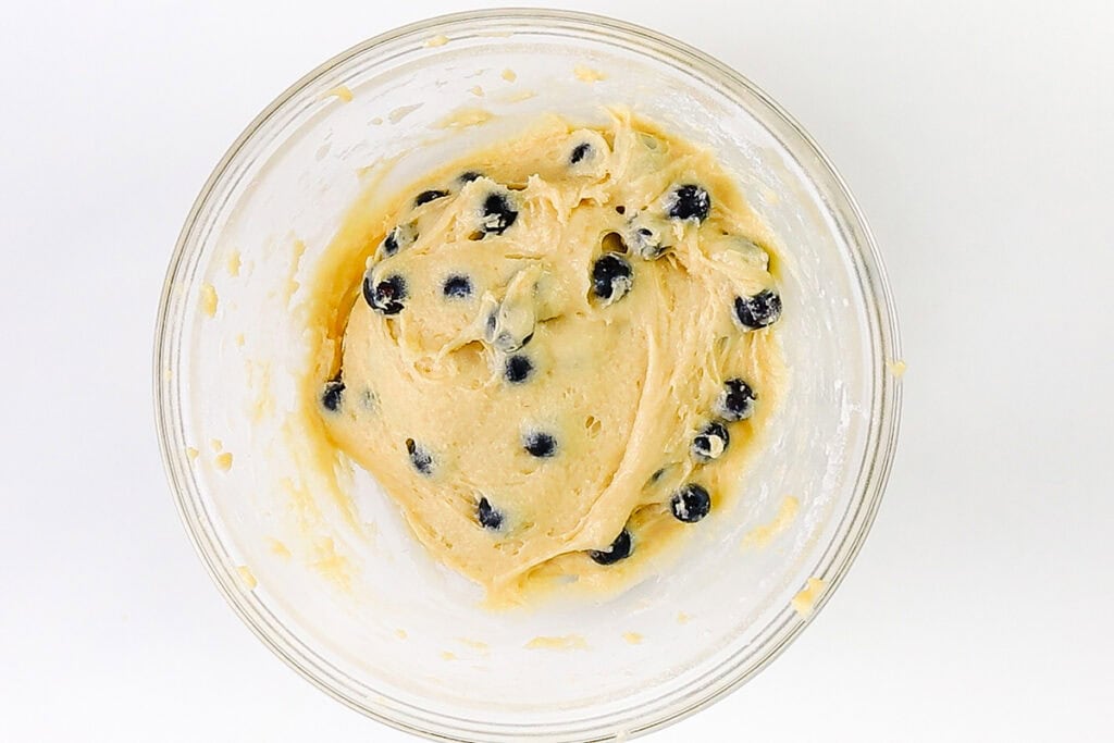 A glass bowl filled with thick, creamy batter mixed with fresh blueberries sits on a white surface, ready to become delicious Blueberry Crumb Cake Muffins.
