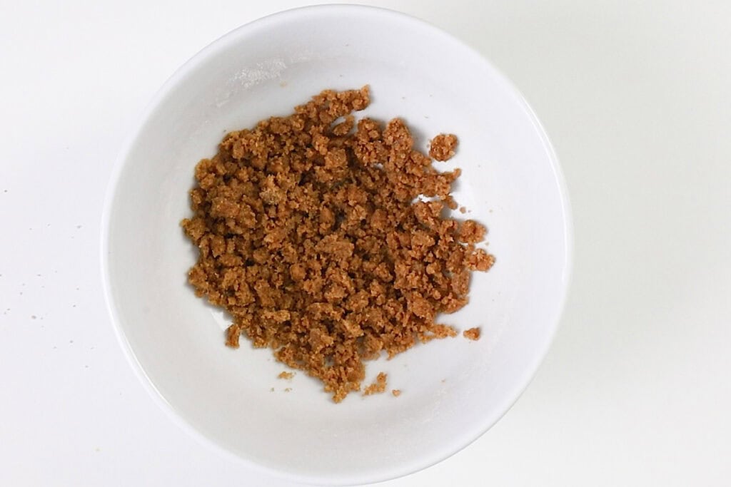 A white bowl containing a mixture of brown, crumbly granules, resembling the topping used for Blueberry Crumb Cake Muffins, sits on a white background.