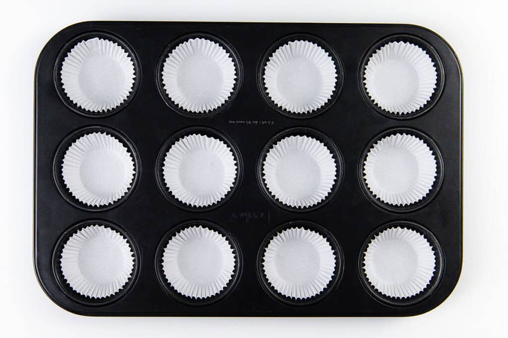 A black muffin tin with twelve cups, each lined with a white paper cupcake liner, ready for baking delicious Blueberry Crumb Cake Muffins, viewed from above on a white background.