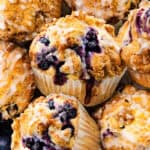A close-up of several Blueberry Crumb Cake Muffins with crumb topping and icing, surrounded by fresh blueberries. The muffins have visible blueberries and a golden-brown, crumbly texture.