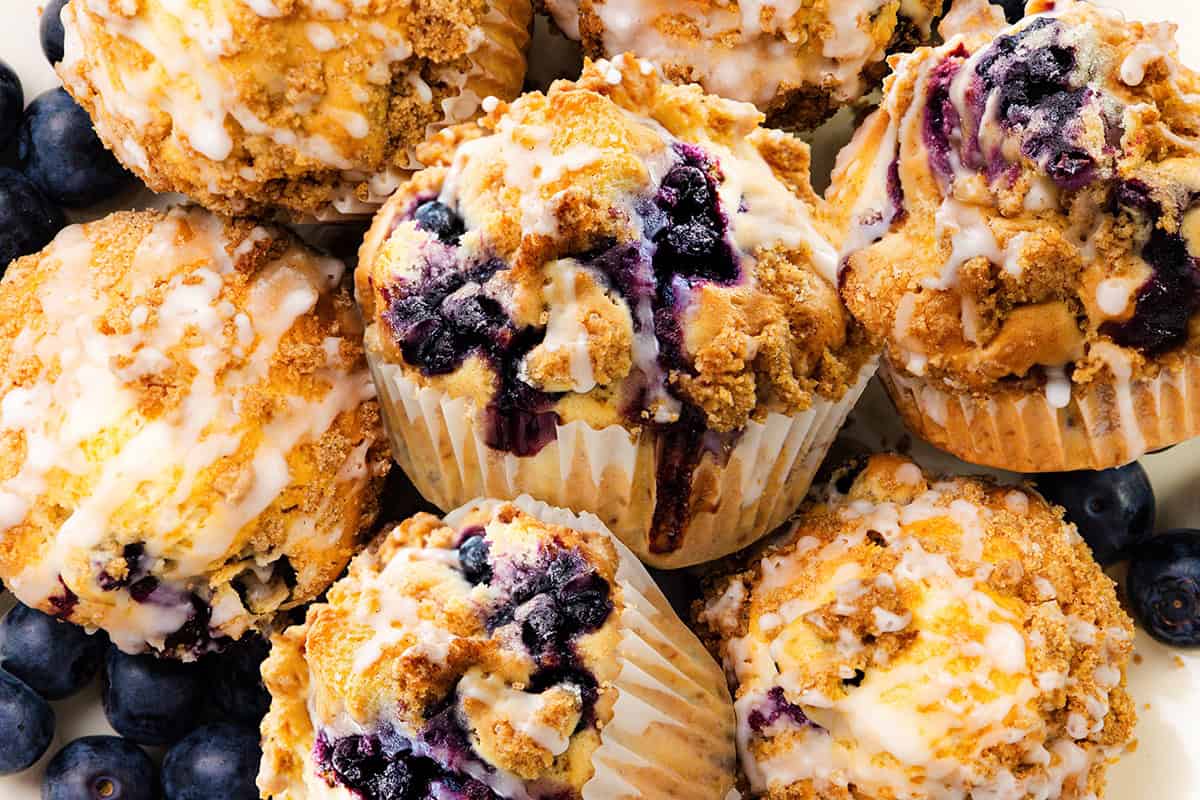 A close-up of several Blueberry Crumb Cake Muffins with crumb topping and icing, surrounded by fresh blueberries. The muffins have visible blueberries and a golden-brown, crumbly texture.