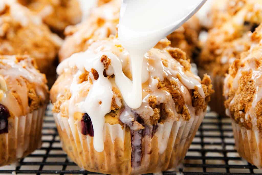 A close-up of Blueberry Crumb Cake Muffins topped with crumbly streusel as white icing is being drizzled over them from a spoon. Other muffins are visible in the background on a cooling rack.