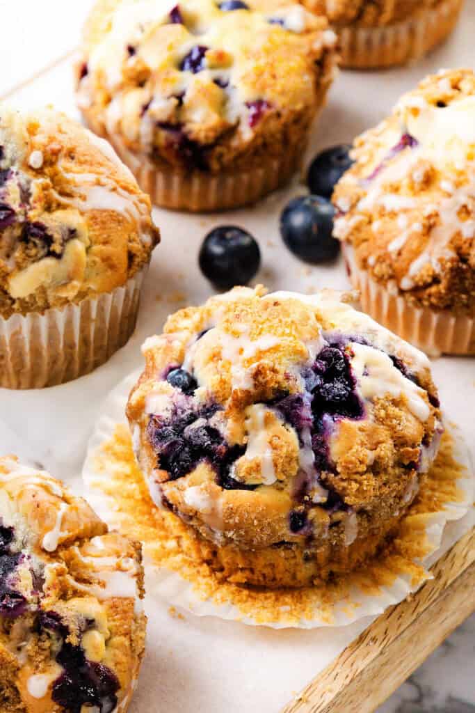 Close-up of Blueberry Crumb Cake Muffins with a crumbly topping and icing drizzle, surrounded by fresh blueberries on a light-colored surface.
