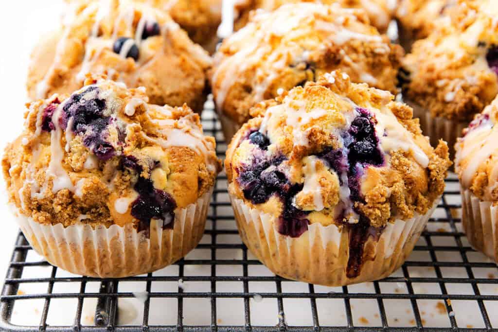 Freshly baked Blueberry Crumb Cake Muffins with crumb topping and icing drizzle are cooling on a wire rack. The muffins are golden brown with visible blueberries and a crunchy, crumbly topping.