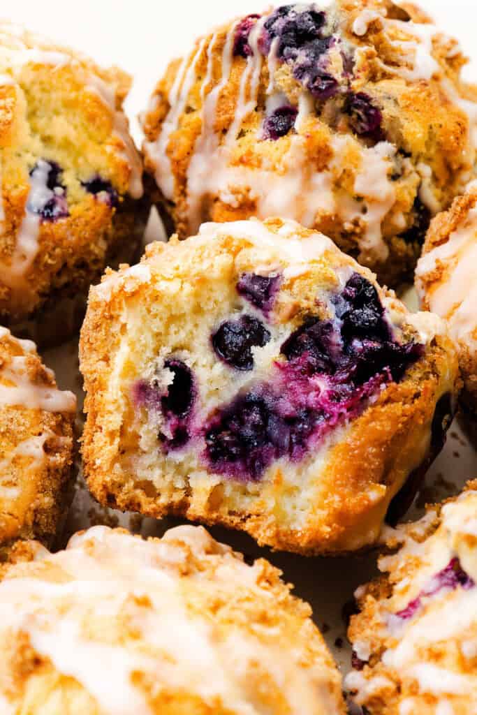 Close-up of a Blueberry Crumb Cake Muffin with a crumbly, golden topping and white glaze, showing juicy blueberries inside. More muffins are partially visible in the background.
