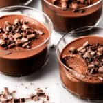 Glass bowls filled with creamy chocolate pots de creme, topped with chocolate shavings. One bowl has a spoonful taken out, revealing the smooth texture inside. The bowls sit on a white surface with scattered chocolate curls.