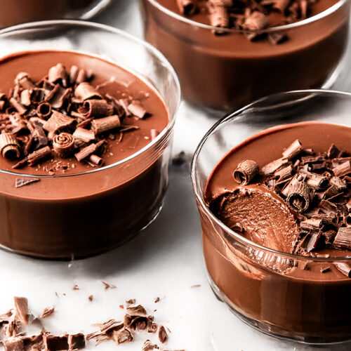 Glass bowls filled with creamy chocolate pots de creme, topped with chocolate shavings. One bowl has a spoonful taken out, revealing the smooth texture inside. The bowls sit on a white surface with scattered chocolate curls.