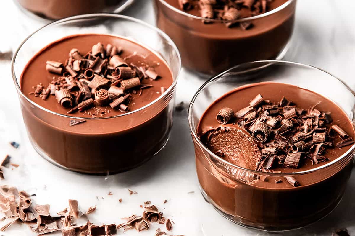 Glass bowls filled with creamy chocolate pots de creme, topped with chocolate shavings. One bowl has a spoonful taken out, revealing the smooth texture inside. The bowls sit on a white surface with scattered chocolate curls.