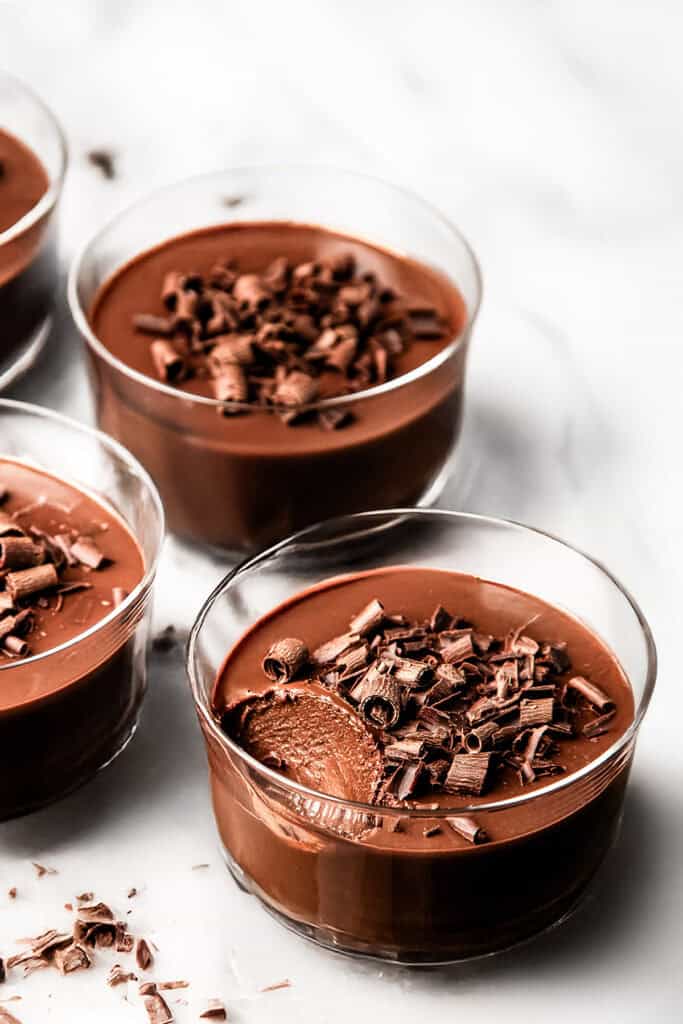 Glass bowls filled with creamy chocolate mousse, reminiscent of chocolate pots de creme, are topped with chocolate shavings. One bowl has a spoonful taken out, revealing the mousse's smooth, rich texture on a white marble surface.
