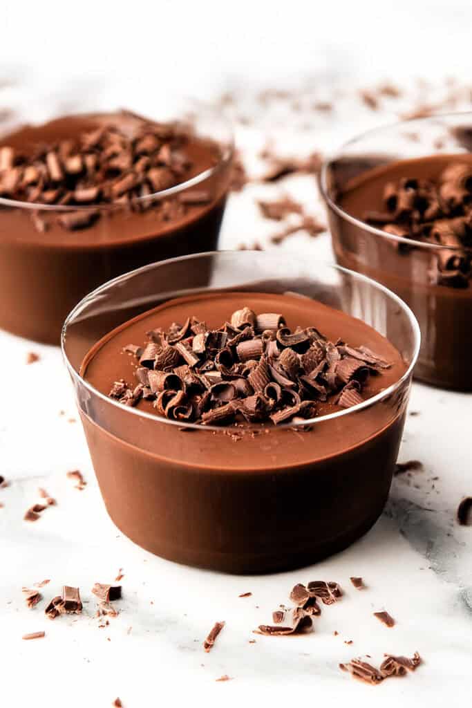 Three glass bowls filled with creamy chocolate pots de creme, each topped with chocolate shavings, are arranged on a white surface with additional chocolate shavings scattered around them.