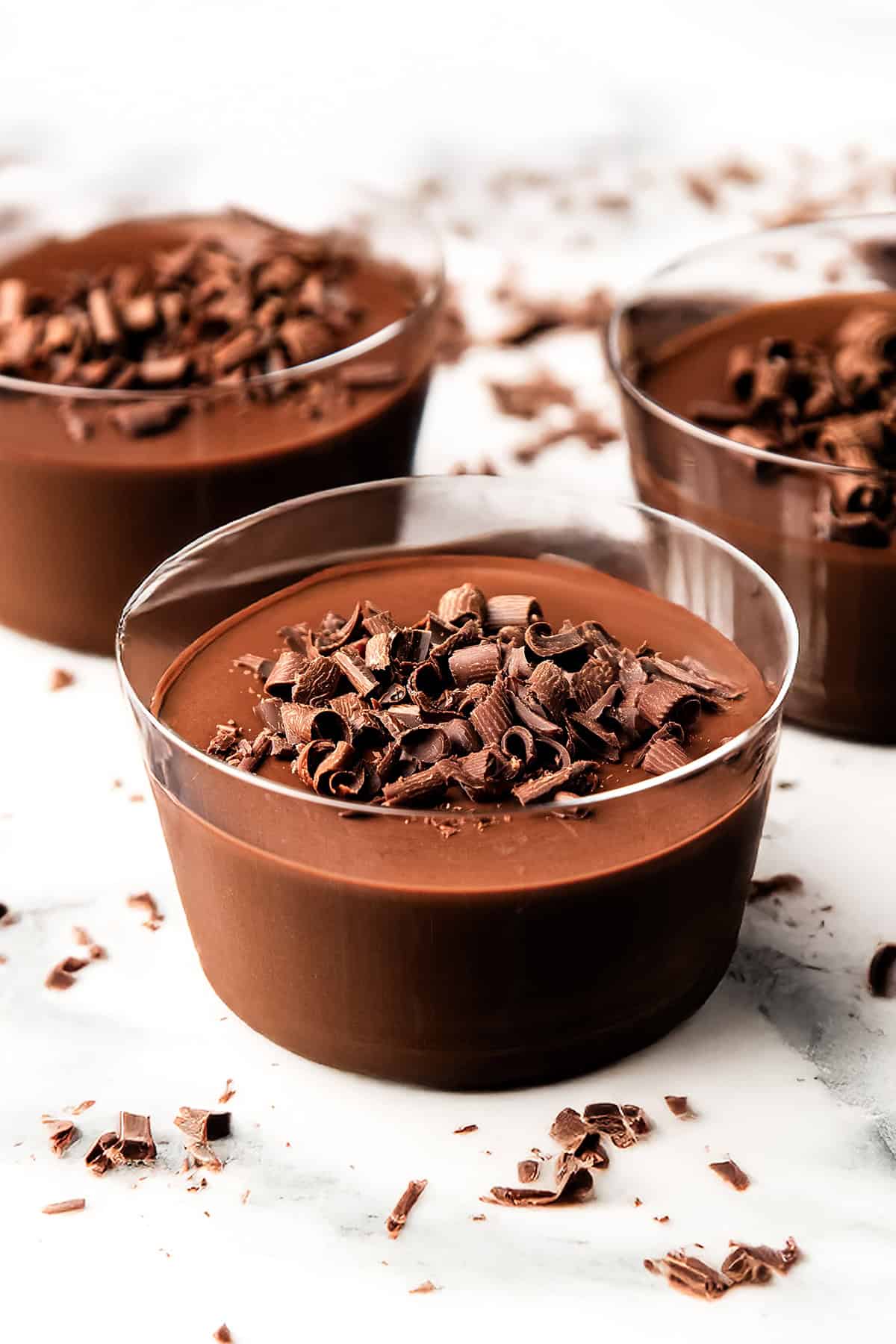 Tiga mangkuk kaca berisi pot de creme coklat krem, masing-masing di atasnya diberi serutan coklat, disusun di atas permukaan putih dengan tambahan serutan coklat tersebar di sekelilingnya.