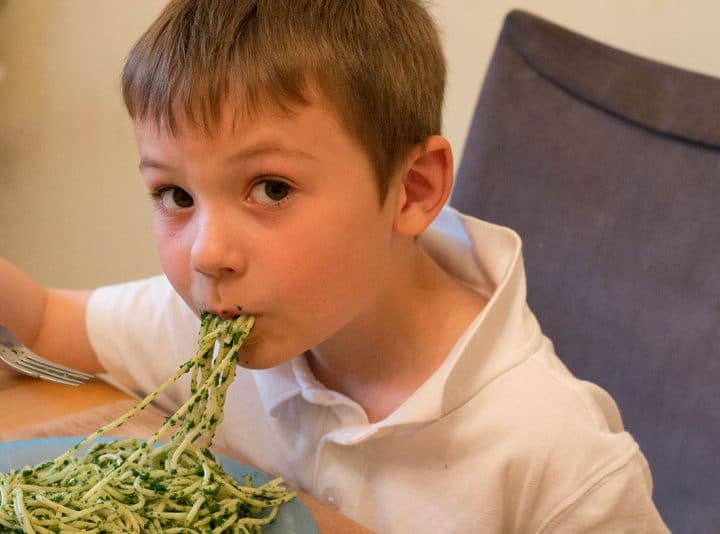 A little boy eating a dish of Spaghetti with Spinach Sauce with strands of spaghetti hanging from his mouth