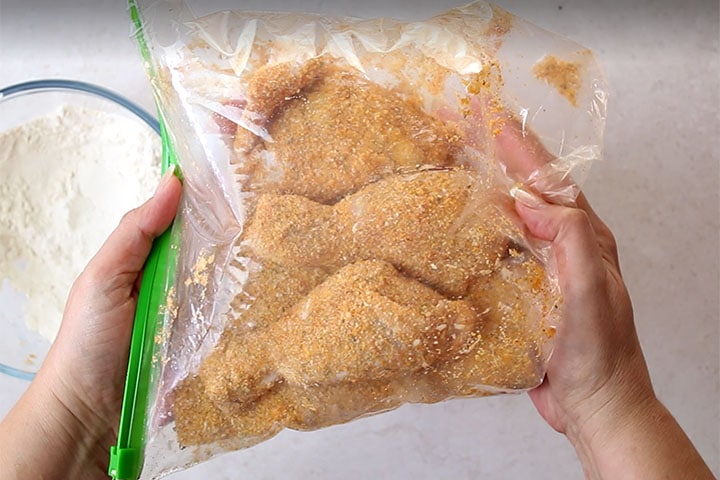 Three pieces of chicken being coated with bread crumbs in a plastic bag.