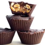 Three homemade Snicker Cups are stacked on top of each other. The top cup is bitten to reveal a filling of caramel, nuts, and possibly nougat. The cups have a dark chocolate outer layer and are displayed against a plain, white background.