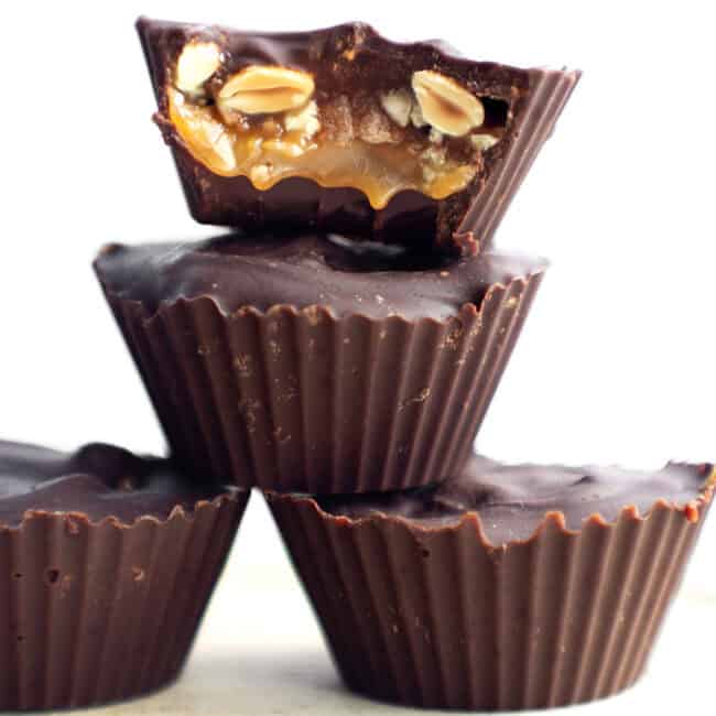 Three homemade Snicker Cups are stacked on top of each other. The top cup is bitten to reveal a filling of caramel, nuts, and possibly nougat. The cups have a dark chocolate outer layer and are displayed against a plain, white background.