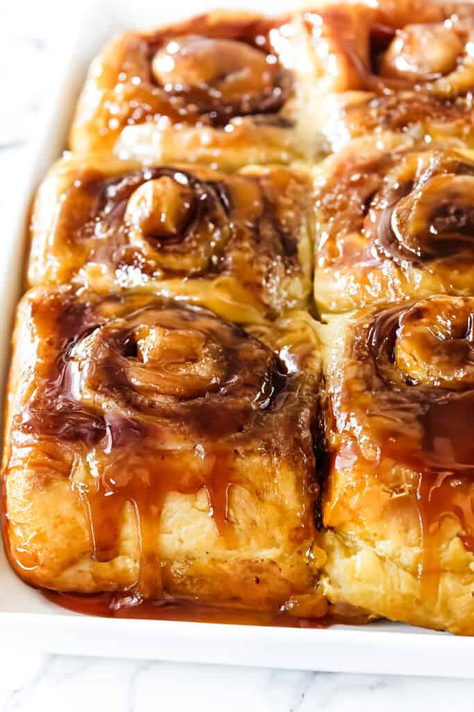 A close-up view of freshly baked sticky buns in a tray, generously coated with a glossy caramel glaze.