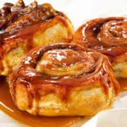 Three freshly made sticky buns covered in caramel sauce and one topped with chopped pecans.