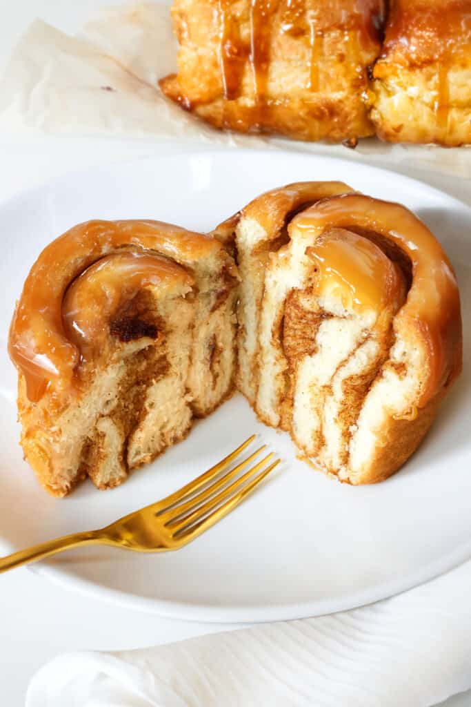 A plate with a sticky bun cut in half showing its cinnamon-swirled interior, drizzled with caramel icing, alongside a gold fork.