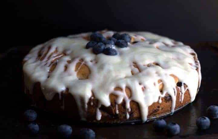 A blueberry cake with a cream cheese icing drizzled all over it topped with fresh blueberries. 