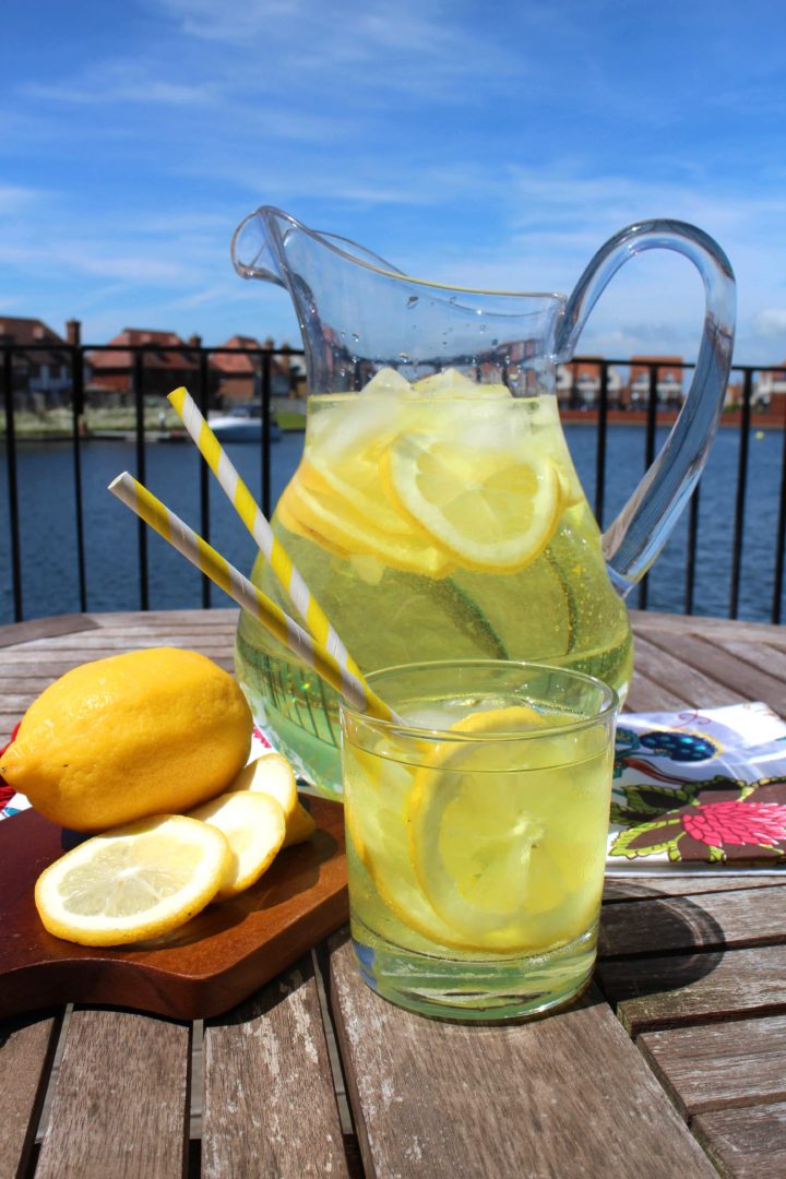 Erren's Hard Lemonade in a glass with a jug and lemons cut up on a chopping board, with a harbour view behind