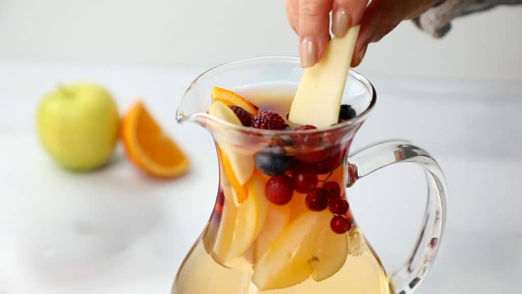 A hand places an apple slice into a glass pitcher filled with sliced fruits and a light-colored beverage, capturing the refreshing preparation of White Sangria. An apple and orange wedges are blurred in the background.