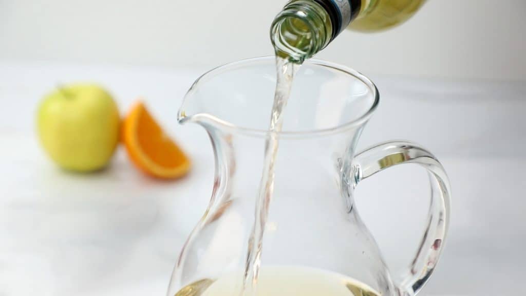 A bottle pours a clear liquid into a glass pitcher, hinting at the refreshing preparation of White Sangria. In the blurred background, there is a green apple and orange slices on a white surface.