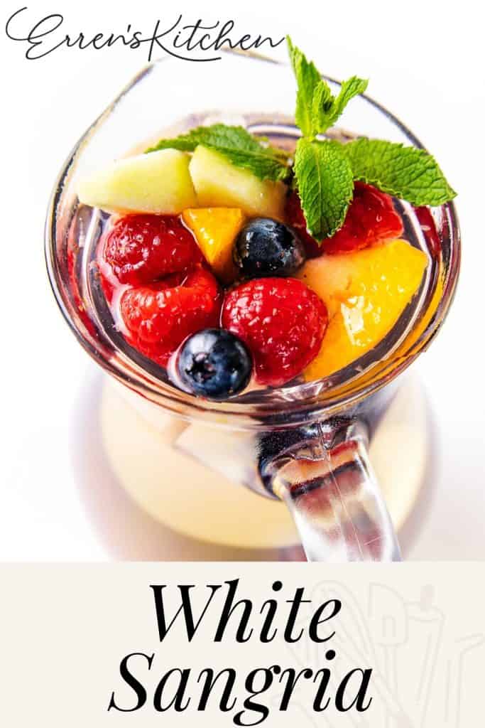 A glass pitcher filled with refreshing White Sangria, garnished with raspberries, blueberries, apple slices, peach pieces, and a mint sprig. The text reads “Erren’s Kitchen White Sangria.”.
