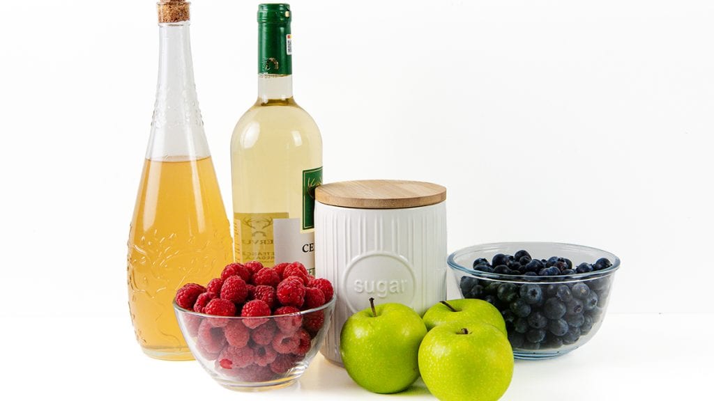 A bottle of white wine, a bottle of white wine perfect for crafting White Sangria, a sugar jar, two green apples, a bowl of blueberries, and a bowl of raspberries are arranged on a white background.