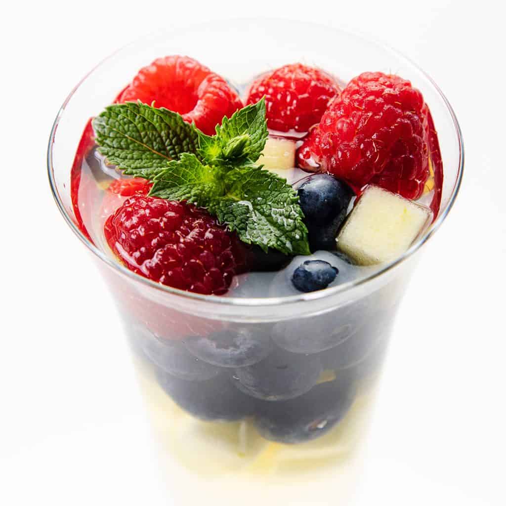 A glass filled with a layered fruit salad, including raspberries, blueberries, apple cubes, and a sprig of fresh mint on top—reminiscent of the vibrant ingredients often found in a refreshing White Sangria—stands against a white background.