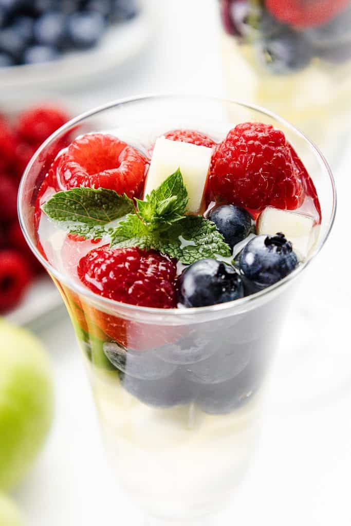 A glass filled with blueberries, raspberries, white fruit cubes, fresh mint leaves, and clear liquid—perfect for showcasing a refreshing White Sangria. Another dish with berries is blurred in the background.