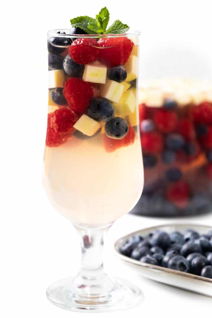 A glass of light-colored White Sangria filled with diced apples, blueberries, and raspberries, garnished with a mint sprig. A tray of blueberries and a container of mixed fruit are visible in the background.