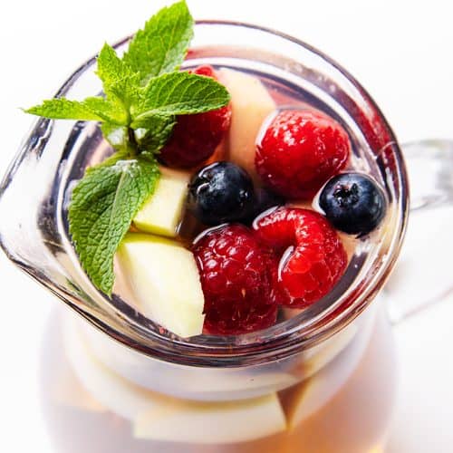 A glass pitcher filled with refreshing White Sangria, topped with raspberries, blueberries, apple slices, and a sprig of fresh mint, shot from above on a white background.