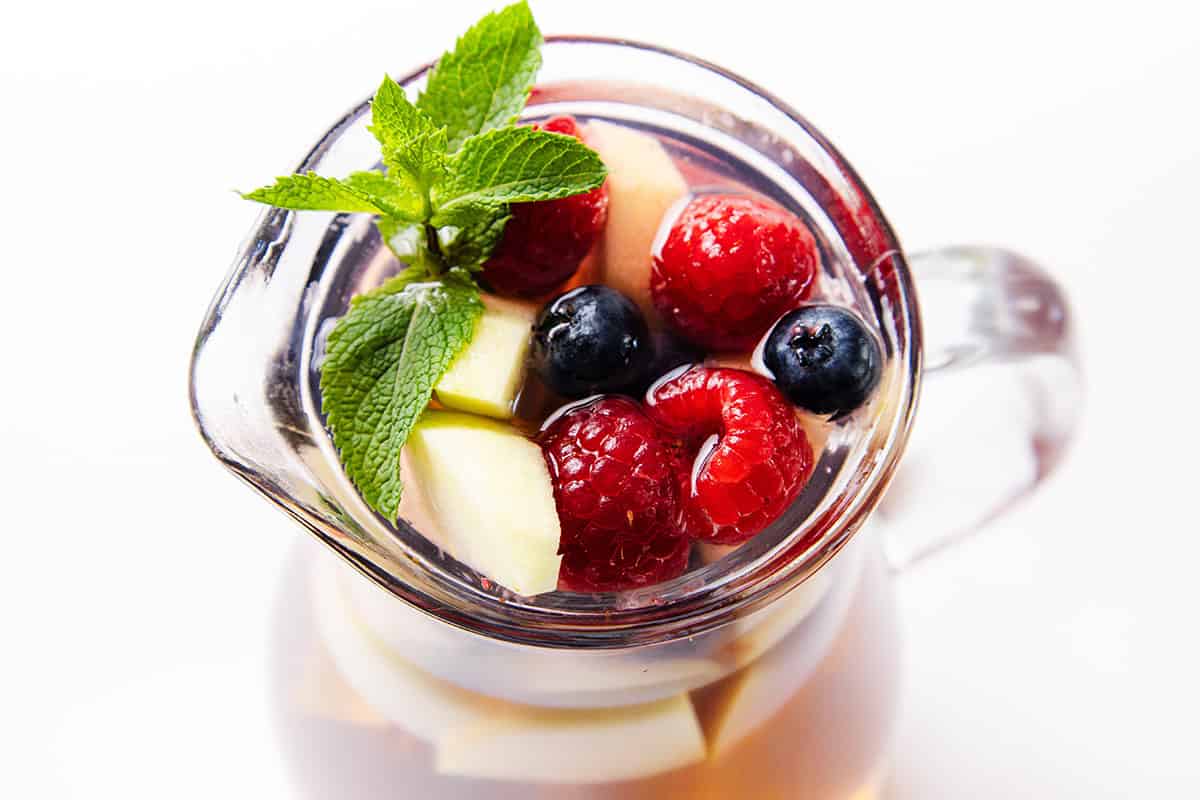 A glass pitcher filled with refreshing White Sangria, topped with raspberries, blueberries, apple slices, and a sprig of fresh mint, shot from above on a white background.