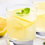 a glass of Homemade Hard Lemonade garnished with lemon slices and a sprig of mint with lemons and another glass in the background