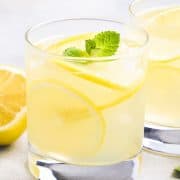 a glass of Homemade Hard Lemonade garnished with lemon slices and a sprig of mint with lemons and another glass in the background