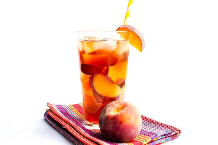 A glass of Peach Iced Tea with ice and a straw and a fresh wedge of peach on the glass