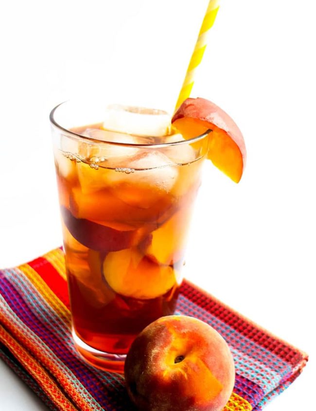 A glass of Sweet Peach Iced Tea with ice and a straw and a fresh wedge of peach on the glass