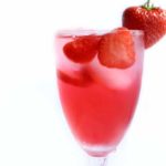 A close up of a glass of strawberry cocktail with strawberries floating in it and a strawberry pushed onto the rim of the glass
