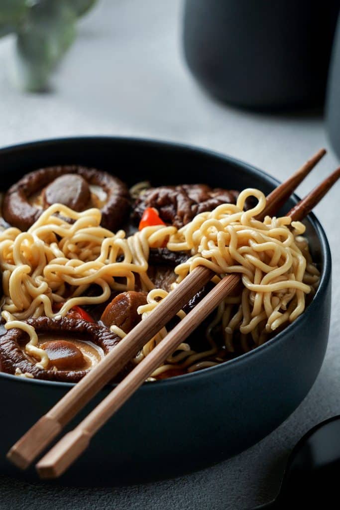 A bowl of chinese noodles with mushrooms and chopsticks ready to serve.