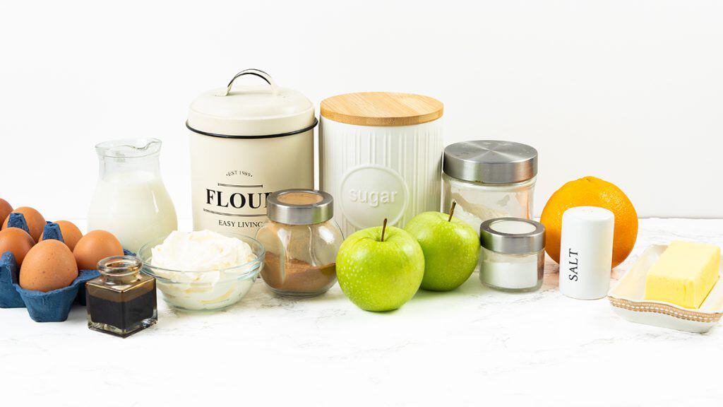 A variety of baking ingredients, perfect for making Apple Cheesecake Muffins, including eggs, milk, flour, sugar, apples, butter, cream, cinnamon, vanilla, an orange, and salt are arranged on a white surface.