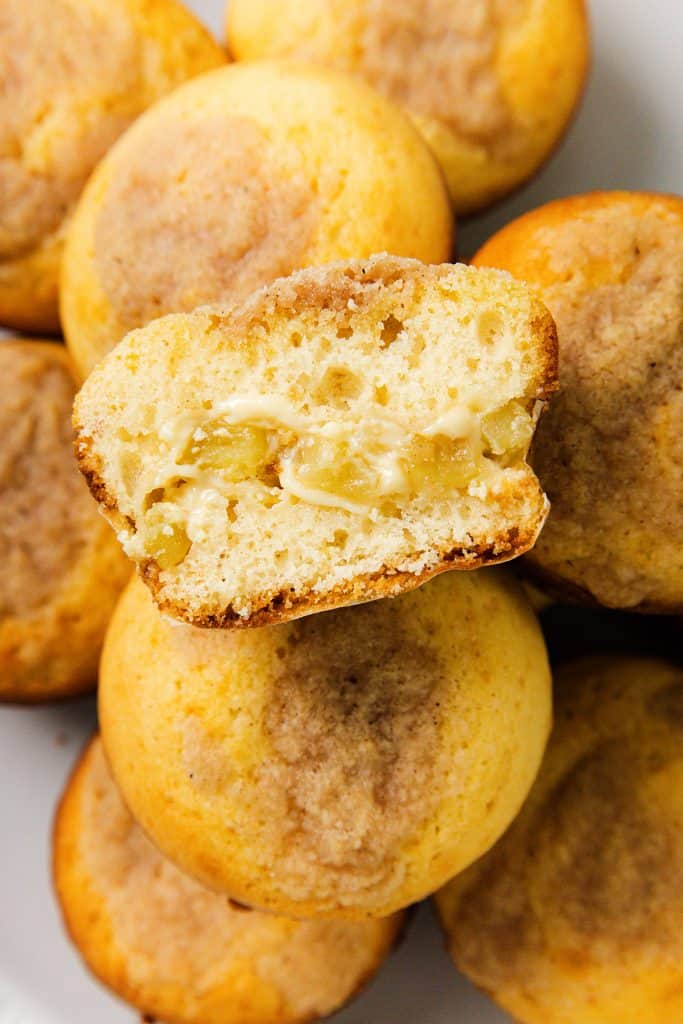 A close-up of a plate of Apple Cheesecake Muffins, with one muffin cut in half, revealing a gooey, creamy filling inside. The muffins are golden brown with a light dusting of cinnamon sugar on top.
