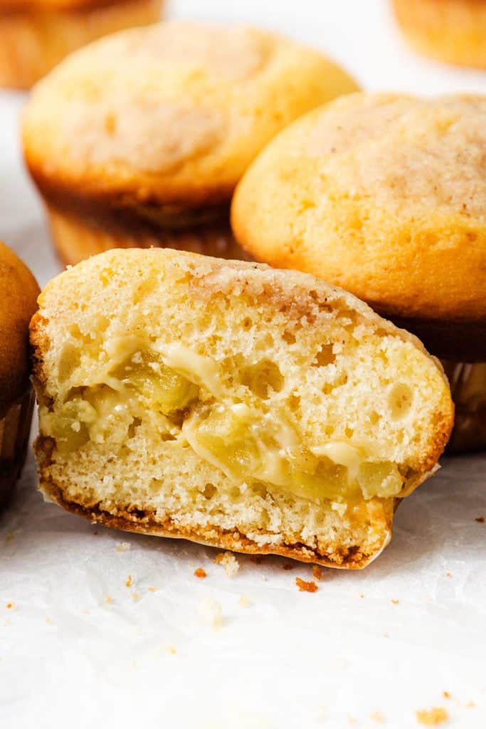 A close-up of a sliced Apple Cheesecake Muffin with a gooey apple filling, showing its soft, moist texture. Whole Apple Cheesecake Muffins are visible in the background on a white surface.