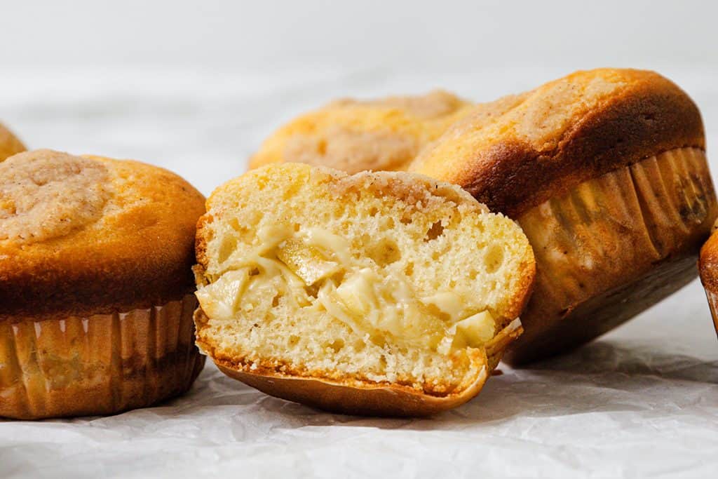 A close-up of Apple Cheesecake Muffins, one cut in half to reveal a melty cheese filling inside. The muffins have a golden-brown top and are set on a crumpled white surface.