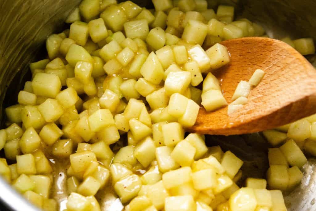 Diced apples being stirred with a wooden spoon in a pot, coated in a glossy sauce, likely being prepared for an apple cheesecake muffins filling or other delectable desserts.